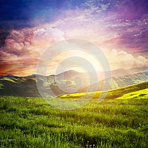 Fairtytale landscape with green grass, mountains, sunset fantastic sky.