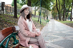 Fall concept - autumn woman drinking coffee on park bench under fall foliage. Beautiful young modern woman smiling happy and