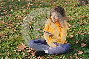 Fall mood. Young girl in yellow sweater with smartphone sitting on autumn grass with dry leaves