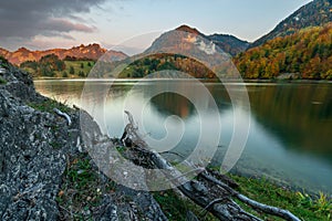 Fall scene taken at sunset of a calm lake surrounded by mountains and a deciduous forest with strong fall colors. The last reddish