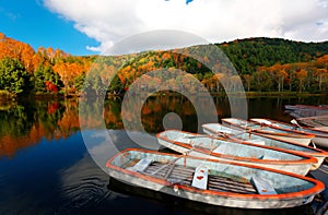 Fall scenery of row boats parking at a wooden dock by Lake Kido Ike & colorful forests on the hillside reflected in the water