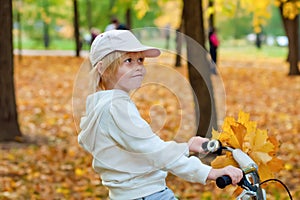 Fall season. Cute blond little caucasian girl riding a bike with yellow autumn maple leaves in a park. Happy childhood