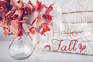Fallen leaves in a vase with pile of white knitted woolen clothes with Hello Fall wording