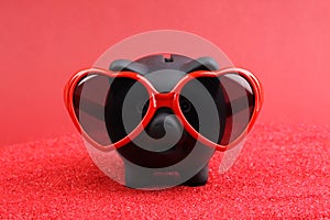 Fallen in love black piggy bank with red heart sunglasses standing on red sand in front of red background