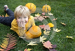 among the fallen multi-colored leaves and pumpkins on the grass lies a cute boy