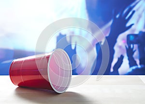 Fallen plastic red party cup on its side on a table. Nightclub or disco full of people dancing on the dance floor.