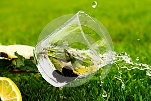 A falling glass of sparkling water with lemon, blackcurrant and mint on grass background. Water flows from a glass.