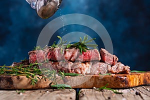 Falling salt on medium rare grilled beef steak ribeye with rosemary on wooden cutting board on dark blue background. Appetizing