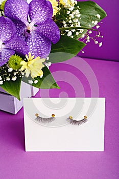 False eyelashes and pink flowers composition on purple background. Beauty products, cosmetics for eyes make up, eyelash extensions