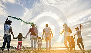 Families friends having fun on the beach with smoke bombs and firework at sunset - Diverse culture sons and parents playing