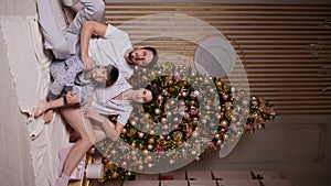 family with a baby boy in pajamas is sitting at the Christmas tree in the new year at home. Vertical. people are looking