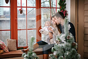 Family with baby boy sitting near the window at home decorated for Christmas