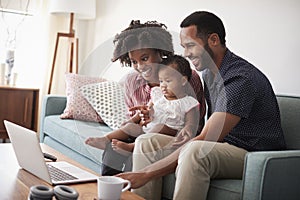 Family With Baby Daughter Sitting On Sofa At Home Looking At Laptop Computer