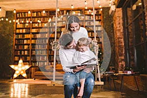 Family With Baby Relaxing On home Swing Seat in cozy dark living room on Christmas eve. Family having fun on seesaw. Winter