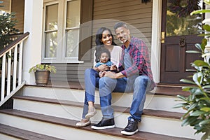 Family With Baby Son Sit On Steps Leading Up To Porch Of Home