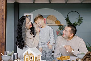 A family bakes a gingerbread house for Christmas with sweets, a mother puts icing on her son`s nose, they laugh