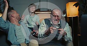 Family bonding time. Happy excited father, teen son and senior grandfather have fun playing console videogames at home.