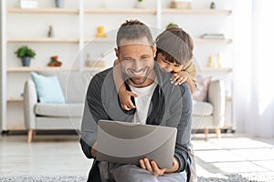 Family business. Smiling man sitting on floor at home and working on laptop, curious little son embracing him