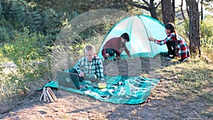 Family camping. Two girls pitch tent and one boy watching show on the laptop, sitting on the plaid. Wanderlust discovery.