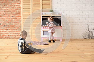 Family and children concept. Toddler boy playing on floor and cute little girl eating sweets at play kitchen in big empty room