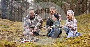 family with children grilling sausages on wooden skewers over a bonfire while camping in forest. nature adventure, bonding