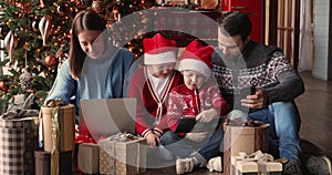 Family with children using gadgets sitting near decorated Christmas tree