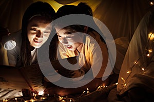 Family concept. Mother and child daughter reading book with flashlight together in children tent before bedtime. Happy mother read