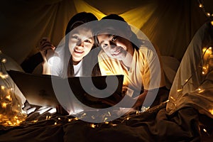 Family concept. Mother and child daughter reading book with flashlight together in children tent before bedtime. Happy mother read