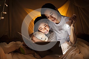 Family concept. Mother and child daughter reading book with flashlight together in children tent before bedtime. Happy mother read