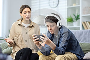 Family conflict quarrel, woman mother quarrels with son, teenage boy in headphones ignores woman and plays video games