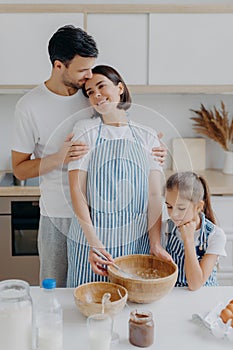 Family cook meal at home. Husband and wife embrace with love, small girl poses near, prepare delicious dinner, use milk, chocolate