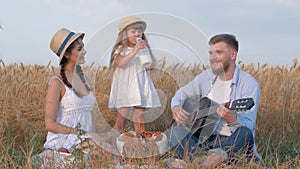 Family in countryside picnic, little girl drinks milk from bottle during outings with her young mother and happy dad