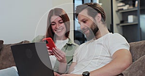 Family couple uses a laptop computer and smartphone, while relaxing on couch of their apartment in the living room. Man