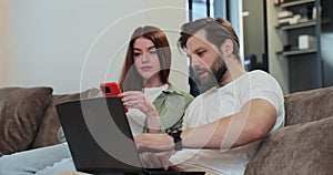 Family couple uses a laptop computer and smartphone, while relaxing on couch of their apartment in the living room. Man