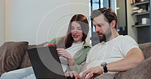 Family couple uses a laptop computer and smartphone, while relaxing on couch of their apartment in the living room. Man