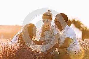 Family day. Happy young mom and dad tickle and hug their little daughter, enjoy relaxing in lavender field at sunset