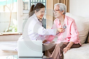 Family doctor or nurse checking smiling senior patient using stethoscope during home visit,young female home caregiver,health
