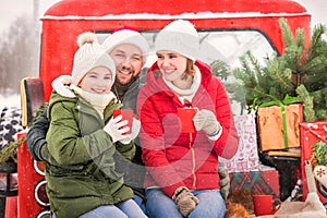 A family drinks tea to warm up in a red car truck with a Christmas tree outside in the snowy winter