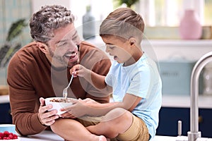 Family With Father In Kitchen With Son Sitting On Counter Eating Breakfast