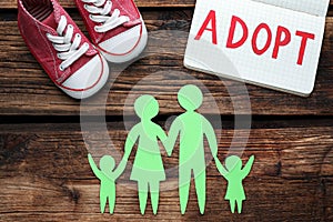 Family figure, child shoes and notebook with word Adopt on wooden table, flat lay