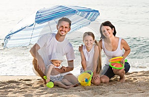 Family of four sitting together under beach umbrella on beach
