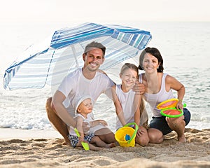 Family of four sitting together under beach umbrella on beach