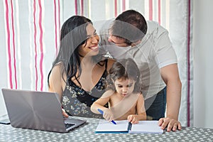 Family gathered at the dining room table, mother at the laptop, father standing hugging them and baby girl painting in a notebook