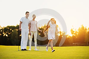 Family on the golf course, happy girl running across the field, her parents are standing with golf clubs behind
