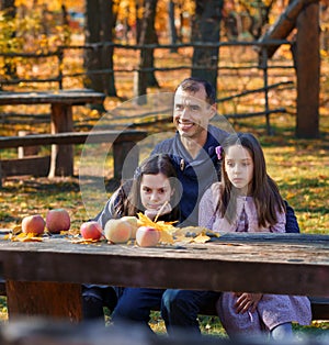 family has picnic in autumn city park, father and two daughters, children and parent sitting together at the table, with apples