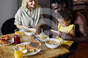 family having breakfast and using smartphone, parents with child in cozy kitchen