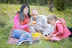 The family is having a picnic in the autumn park. Mom dad and daughter are sitting on the grass in the forest. Parents and
