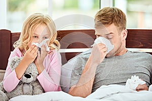 Family health. Young father and his daughter are suffering from flu or cold and runny nose while lying in bed together