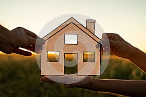 Family holds paper house at sunset, sun shines through window. Hand holding paper cut of house symbols at sunset