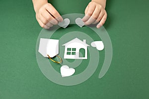 Family home and real estate concept. Close up of child hands holding white paper house and heart on green background.